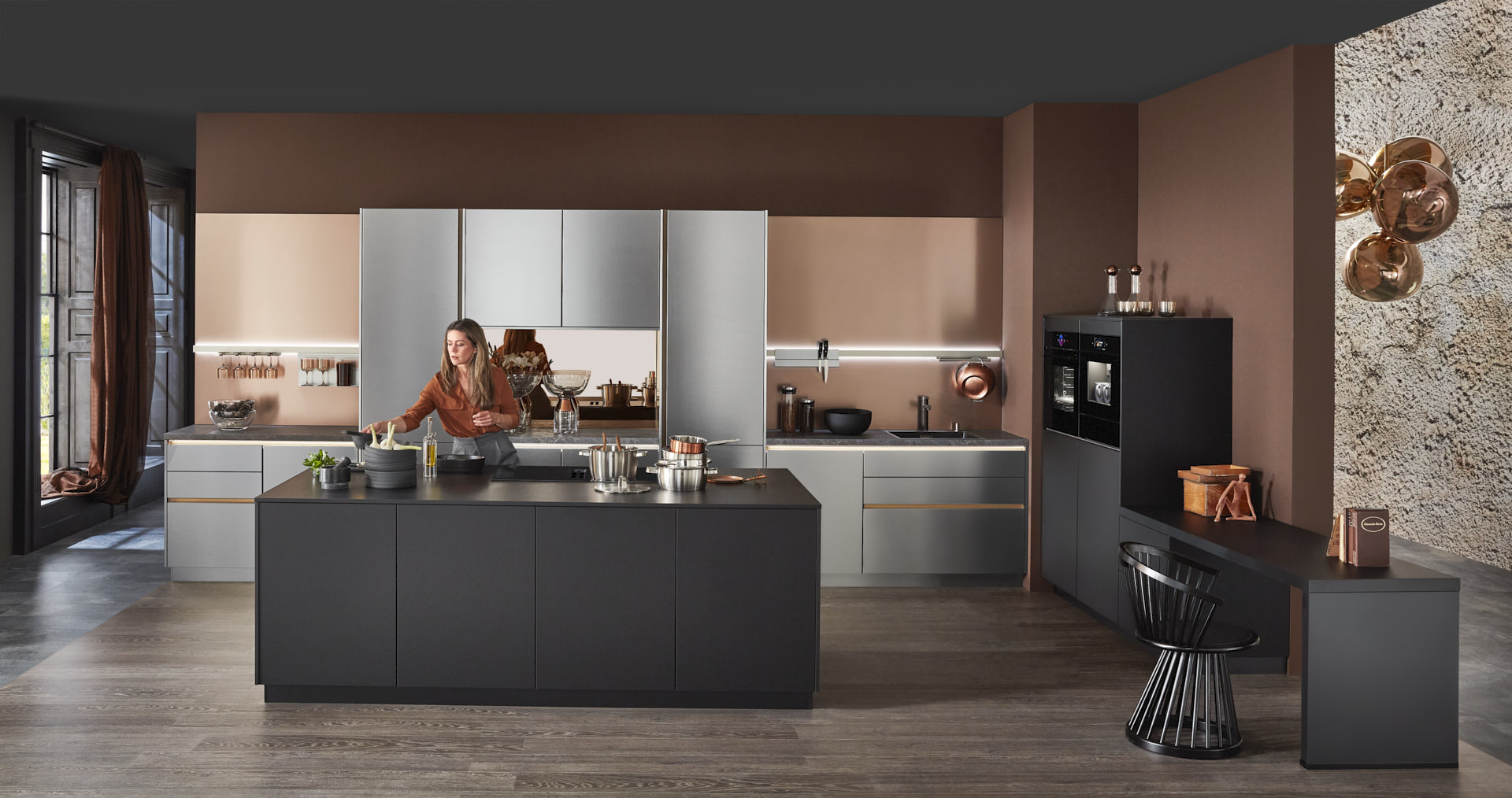 Hauptschuss, Mix aus Keramik und Bronzenachbildung, Kücheninsel mit Bosch Induktionskochfeld und Muldenlüfter, schwarzer Schrank, Edelstahlfronten, Bronze Nischenrückwand, 2 Bosch Einbaubacköfen, Messing Spüle, Model ist hinter Kücheninsel